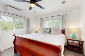 A bedroom with a wooden bed, two bedside tables with lamps, a ceiling fan, and windows with blinds. - Hawaiian Shores Family Retreat near Hot Ponds- Walk to Beach - Dog Friendly (Pahoa)