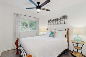 Ein Schlafzimmer mit einem Bett, Deckenventilator, einem Fenster mit Vorhängen, einem Wandbild und einem Nachttisch mit Lampe.