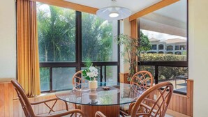Zona pranzo con tavolo in vetro, sedie in vimini e vista su vegetazione ed edifici esterni.