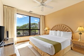 Une chambre d’hôtel avec un lit, une télévision, une fenêtre donnant sur des arbres et un ciel dégagé, et un ventilateur de plafond.