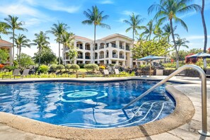 A swimming pool with a clear blue center, surrounded by a sandy edge, with a palm tree and a building with multiple balconies in the background. - *NEW* Top-Floor Waikoloa Escape with Mountain and Golf Views (Waikoloa)