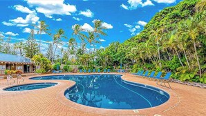 Área de piscina com duas piscinas circulares, cercada por palmeiras e um edifício com uma placa.