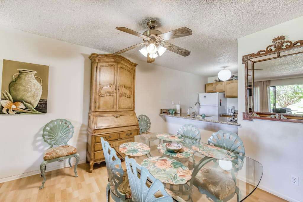 Un comedor con mesa de vidrio, sillas con estampado floral, un aparador de madera y un ventilador de techo.
