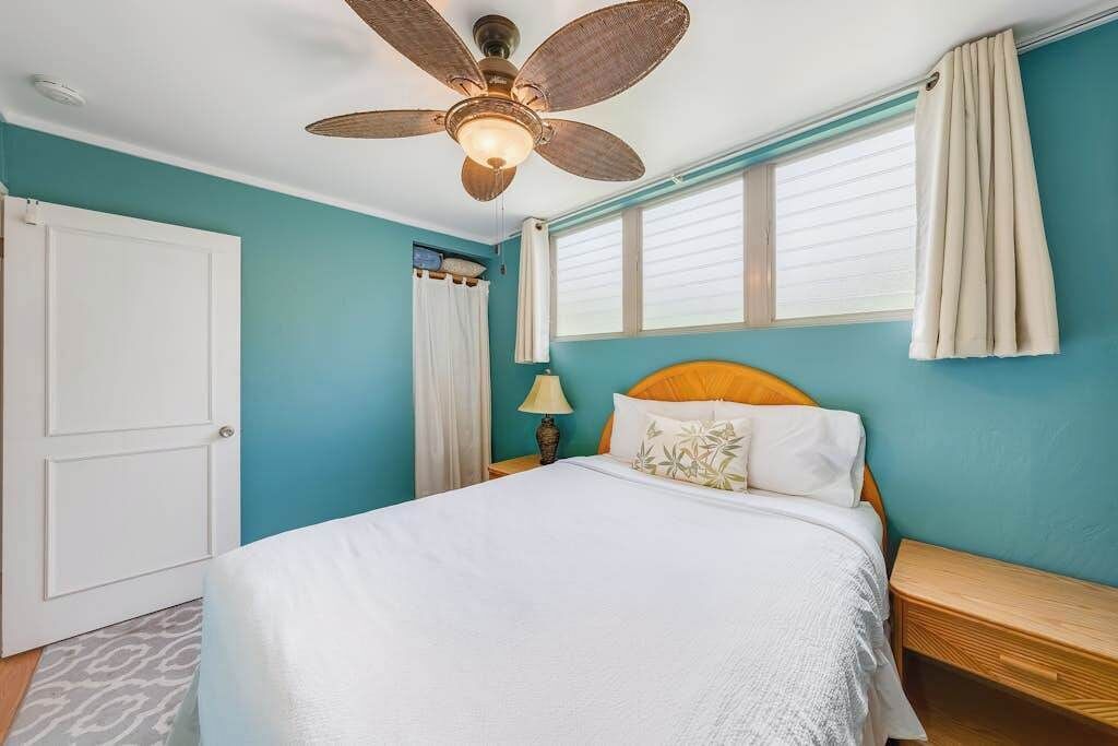 A bedroom with a bed, a ceiling fan, a nightstand, a lamp, and a window with curtains.