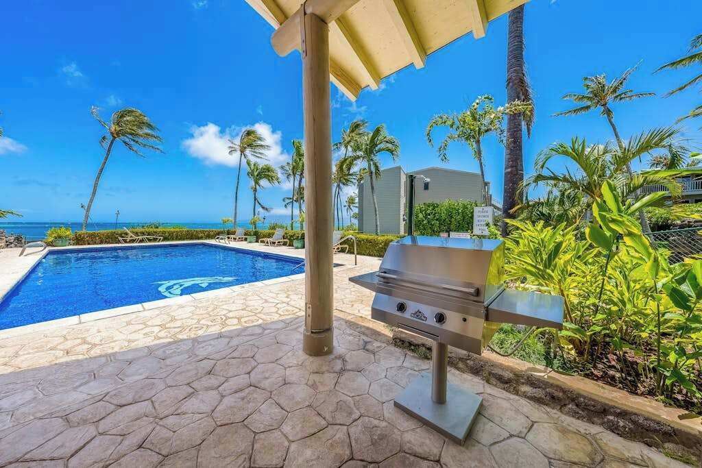 A pool with a view of the ocean, a grill, and palm trees.