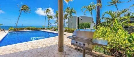 A pool with a view of the ocean, a grill, and palm trees.