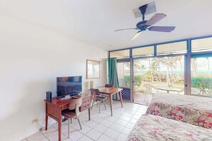 A room with a bed, a desk with a TV, a ceiling fan, and large windows with a view of palm trees.