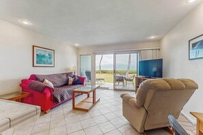 A living room with a red sofa, a beige armchair, a coffee table, a television, and a large sliding glass door leading to an outdoor area. - New Fall $pecial First-Floor Molokai Oceanfront Escape with Pool (Kaunakakai)