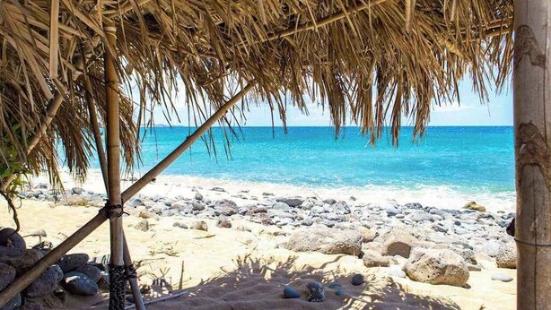 Ein Strand mit einer strohgedeckten Struktur, klarem blauen Wasser und einer felsigen Küste.