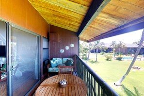 Eine Holzterrasse mit rundem Tisch und einer Couch, mit Blick auf einen Pool und Palmen.