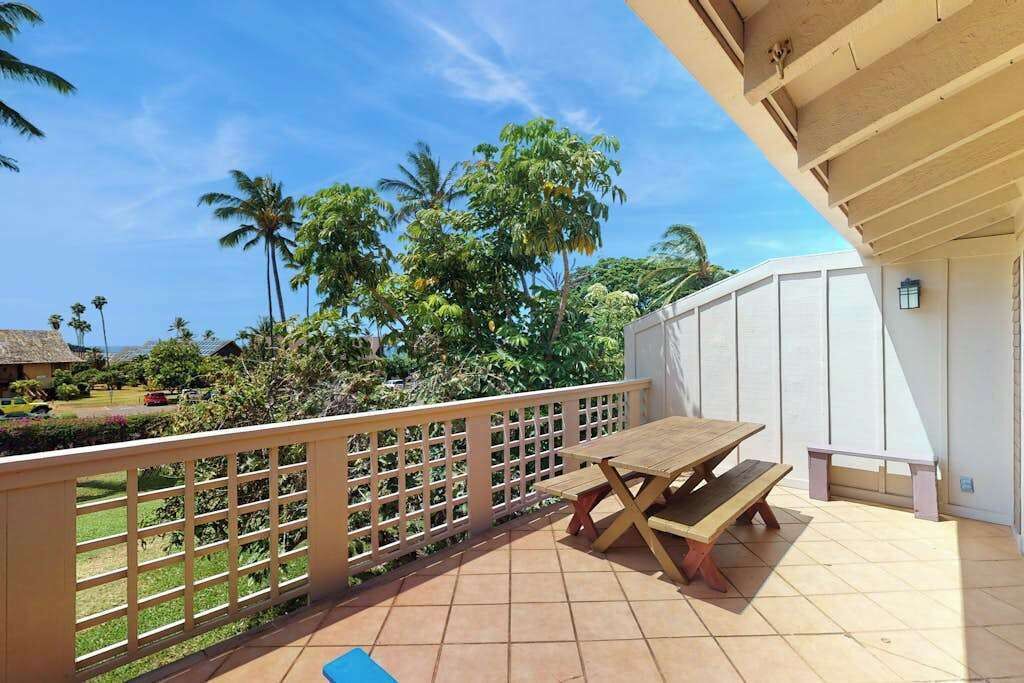 A balcony with a wooden table and bench, a white fence, and a view of palm trees and a house.