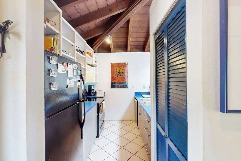 A narrow kitchen with a refrigerator, cabinets, and a window with blinds.