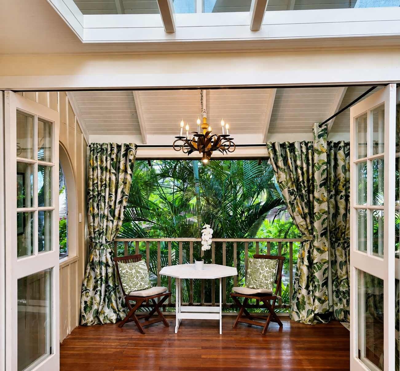 A room with a wooden floor, a round table, two chairs, a chandelier, and a view of greenery through curtains.