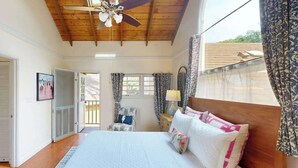 Quarto com teto de madeira, cama com roupa de cama branca, uma cadeira, mesa de cabeceira e janela com cortinas.