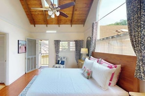 A bedroom with a wooden ceiling, a bed with white bedding, a chair, a nightstand, and a window with curtains. - NEW Fall Sale Colorful Molokai Condo Near Kepuhi Beach with Lanai and Pool (Maunaloa)