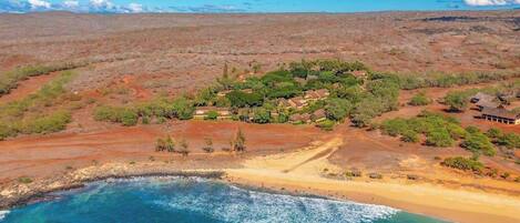 Un paisaje costero con playa, aguas azul turquesa y un pequeño grupo de edificios rodeados de vegetación escasa.