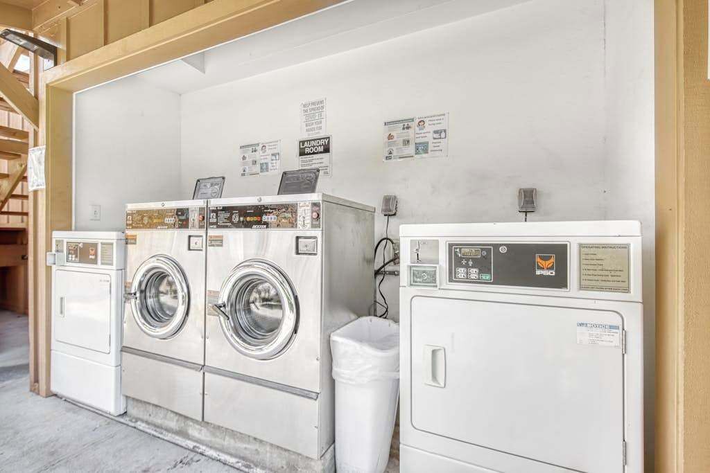 A laundry room with two front-loading washers and a dryer, a trash bin, and informational posters on the wall.