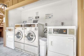 A laundry room with two front-loading washers and a dryer, a trash bin, and informational posters on the wall.