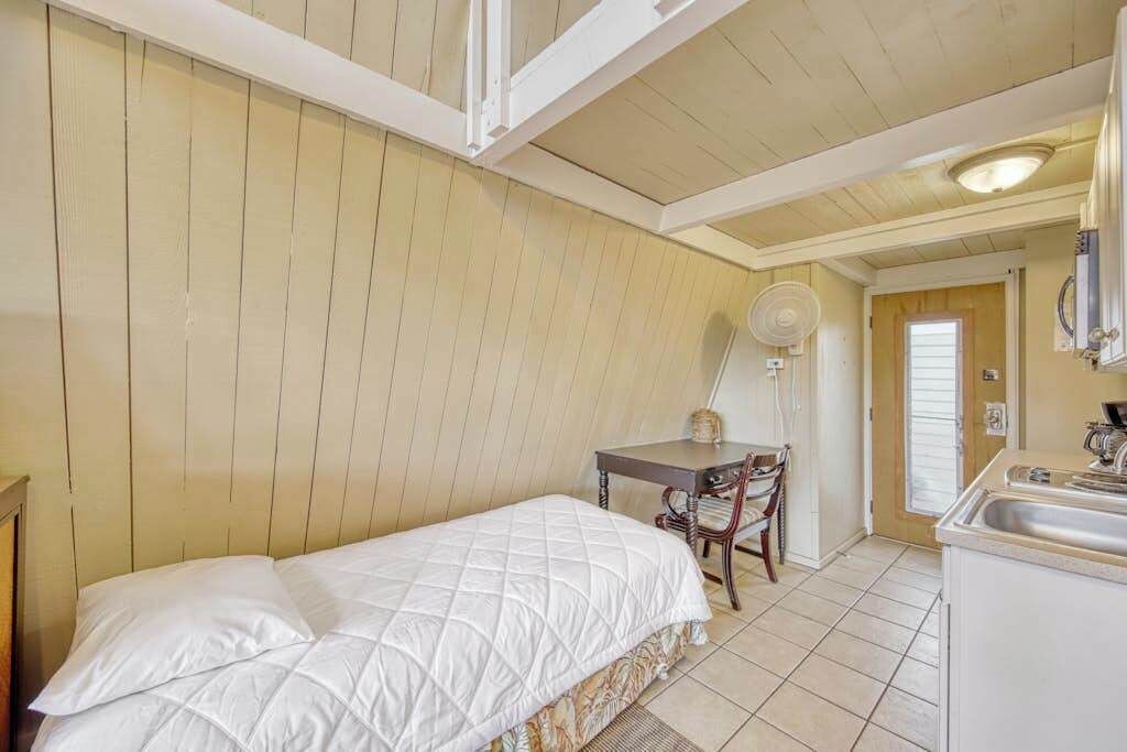 Een kamer met een bed, een bureau, een stoel, een ventilator en een keukenhoek.