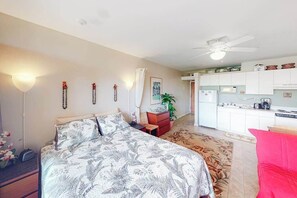 Une chambre à coucher comprenant un lit, un fauteuil rouge, un bureau, une plante, et un coin cuisine avec des placards et des appareils électroménagers.