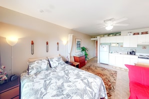 A bedroom with a bed, a nightstand, a red chair, a plant, and a kitchen area with cabinets and appliances.
