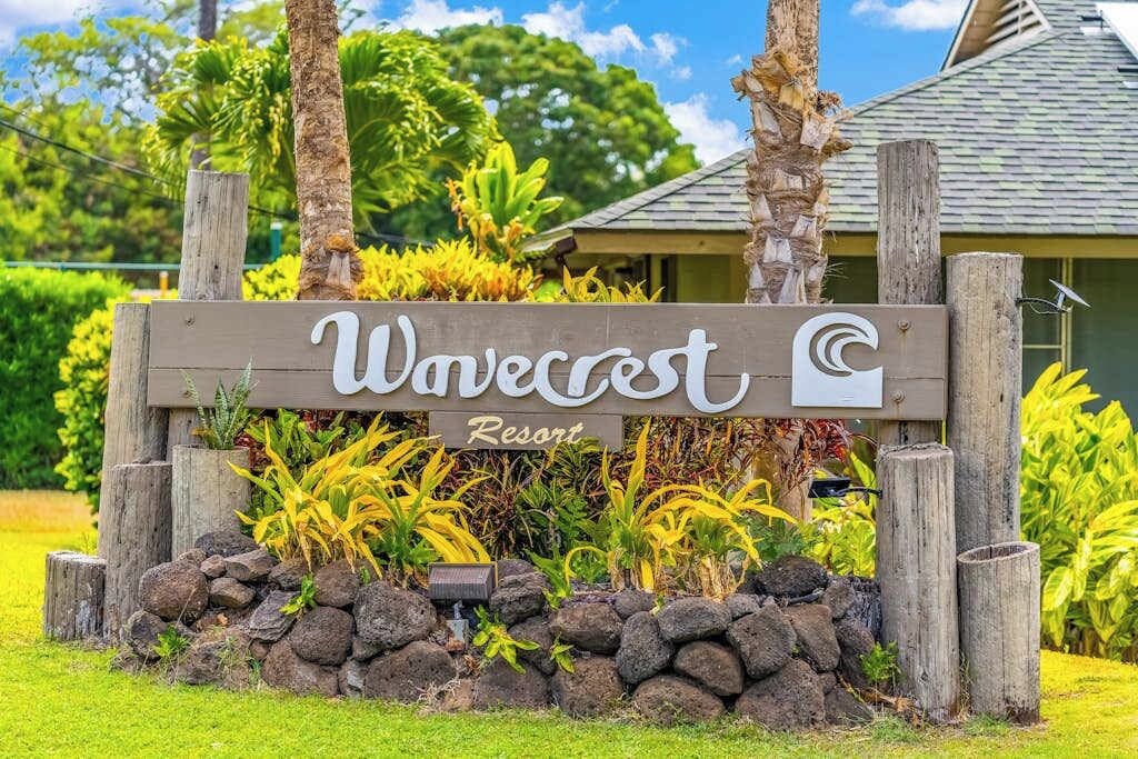 A wooden sign with 'Wavecrest Resort' written on it, surrounded by tropical plants and a stone wall.