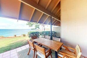 A covered patio with a wooden table and chairs, overlooking a grassy area and the ocean. - NEW Fall Special Oceanfront Molokai Cottage Steps from Kepuhi Beach (Maunaloa)