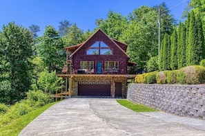 A two-story wooden cabin with a garage, surrounded by lush greenery and a stone wall. - Birchwood Lodge (Pigeon Forge)