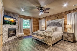 A bedroom with a fireplace, a bed with a tufted headboard, a bench, and a nightstand. - Birchwood Lodge (Pigeon Forge)