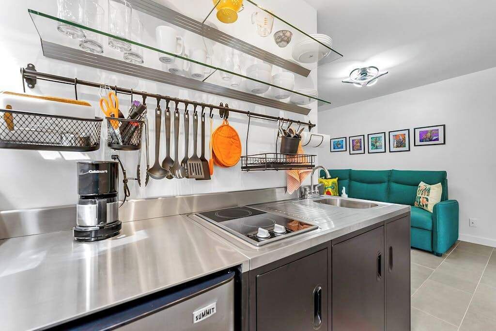 A modern kitchen with stainless steel appliances, a built-in oven, and a coffee maker. There is a teal sofa and a wall-mounted shelf with kitchen utensils.