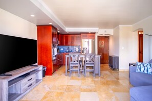 A modern kitchen with red cabinets, a dining area with a wooden table and chairs, a grey sofa, and a flat-screen TV. - New Waikiki City and Mountain Views with Pool and Sauna (Honolulu)