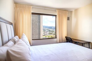 A hotel room with a bed, a desk, and a view of a cityscape through a window with curtains. - New Waikiki City and Mountain Views with Pool and Sauna (Honolulu)