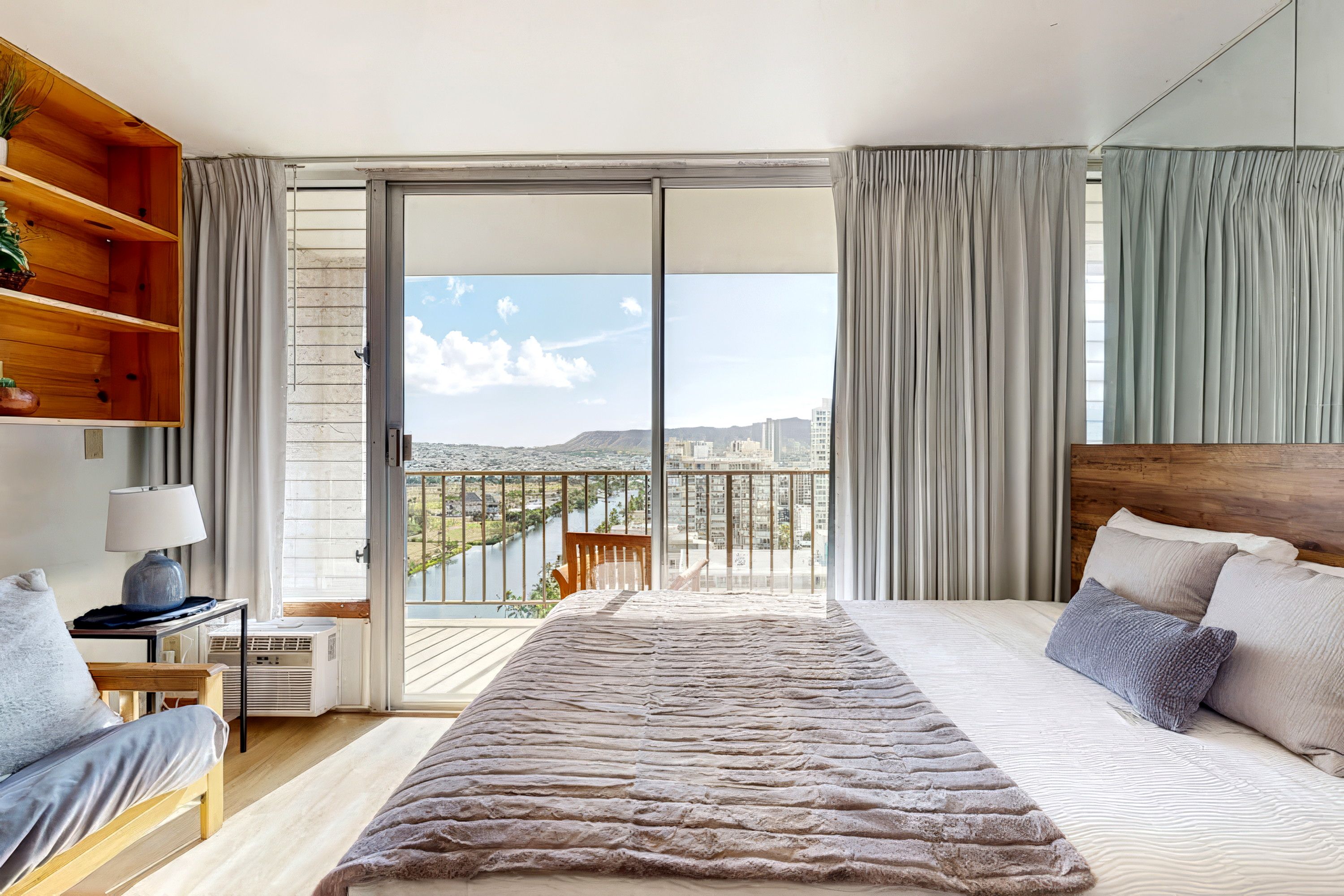 Una camera da letto con un letto, un comodino, una lampada, una finestra con tende e vista sulla città.