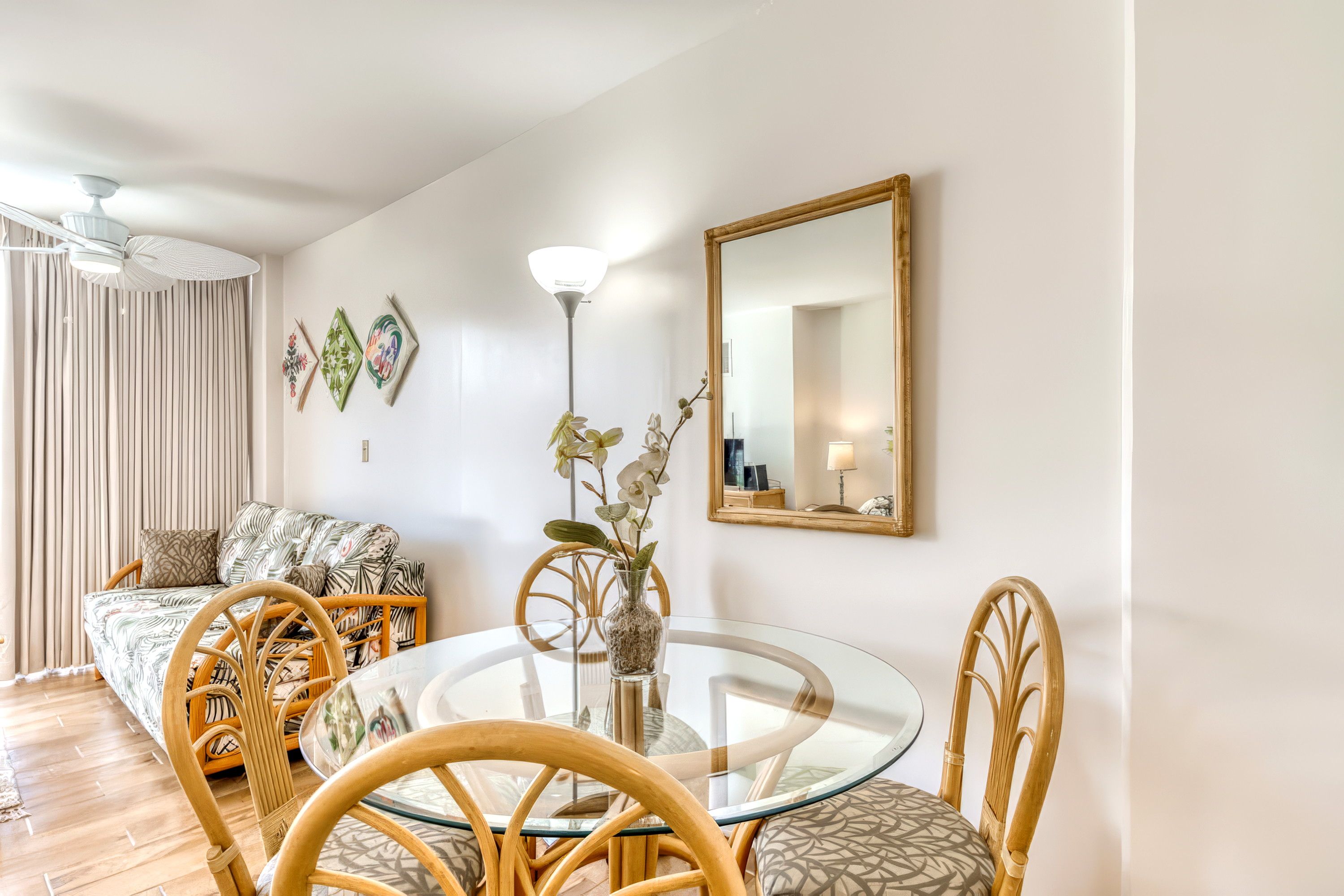 Ein modernes Wohnzimmer mit einem runden Glastisch, Korbstühlen, einer Couch mit Blattmuster, einer Stehlampe und einem Spiegel an der Wand.