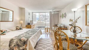 Quarto de hotel com cama, sofá, mesa de jantar com cadeiras, varanda e vista para prédios, e um ventilador de teto.