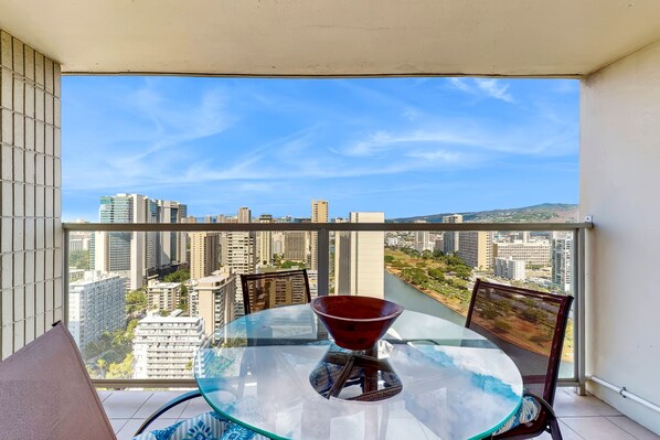 A balcony with a glass table and chairs, overlooking a cityscape with high-rise buildings. - Waikiki Studio High Rise with Lanai and Pool - 30 night minimum (Honolulu)