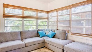 Ein Eckwohnzimmer mit Ecksofa, großen Fenstern mit Jalousien und einem gemusterten Teppich.