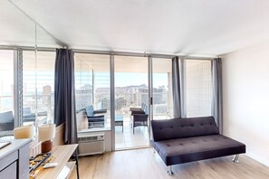 A modern living room with a sofa, a dining table, and a large window offering a city view. - NEW Fall $pecial Waikiki Studio with Diamond Head and Ocean Views (Honolulu)