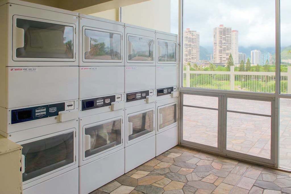 A laundry facility with multiple washing machines, a stone-tiled floor, and a view of a cityscape through a large window.