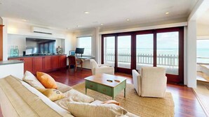 A spacious living area with a sofa, coffee table, and chairs, a kitchen area with cabinets and a TV, and a large window with a view of the ocean. - NEW Historic Tonggs Beach Home with Three Sunset Lanais (Honolulu)