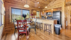 A wooden cabin interior with a dining area, kitchen, and large windows. - Brave Eagle (Gatlinburg)