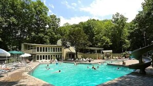A swimming pool with lounge chairs and umbrellas, surrounded by trees and a building with large windows. - Chalet in the Smokies (Gatlinburg)