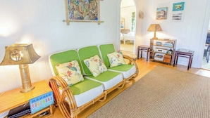 A room with a wicker sofa, a lamp, a small table, and a wall with a painting. - *New* Two Waikiki Cottages with Diamond Head Views | 30 Nights (Honolulu)