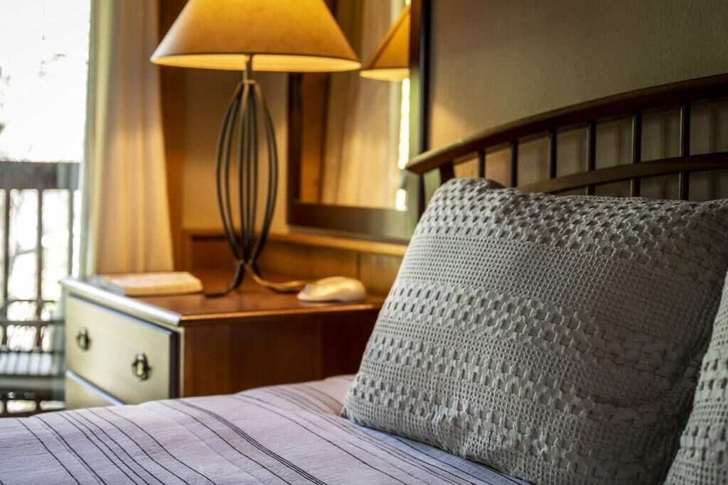 A bed with a striped bedspread, a knitted pillow, a bedside lamp, and a wooden nightstand with a telephone.
