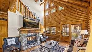 A log cabin interior with a stone fireplace, wooden staircase, and a view of the outdoors. - Cozy Bear Cove (Pigeon Forge)