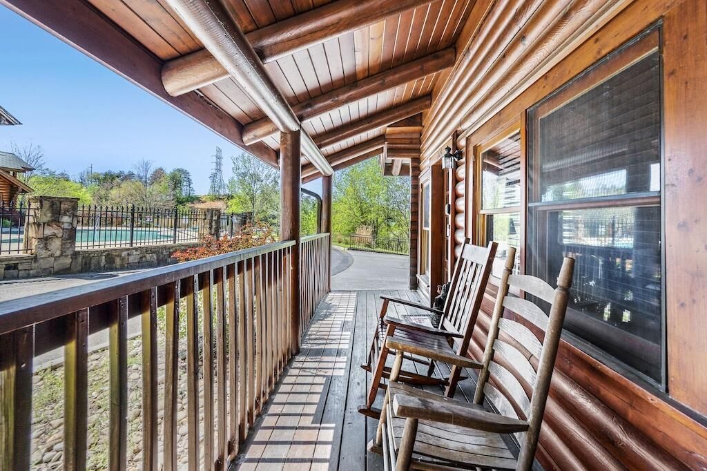 Una terraza de madera con barandilla, bancos de madera y vista a una piscina y árboles.
