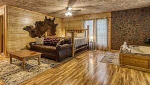 A log cabin interior with a wooden ceiling, a bed, a sofa, a coffee table, a stone wall, and a bathtub. - Cozy Bear Lodge (Sevierville)