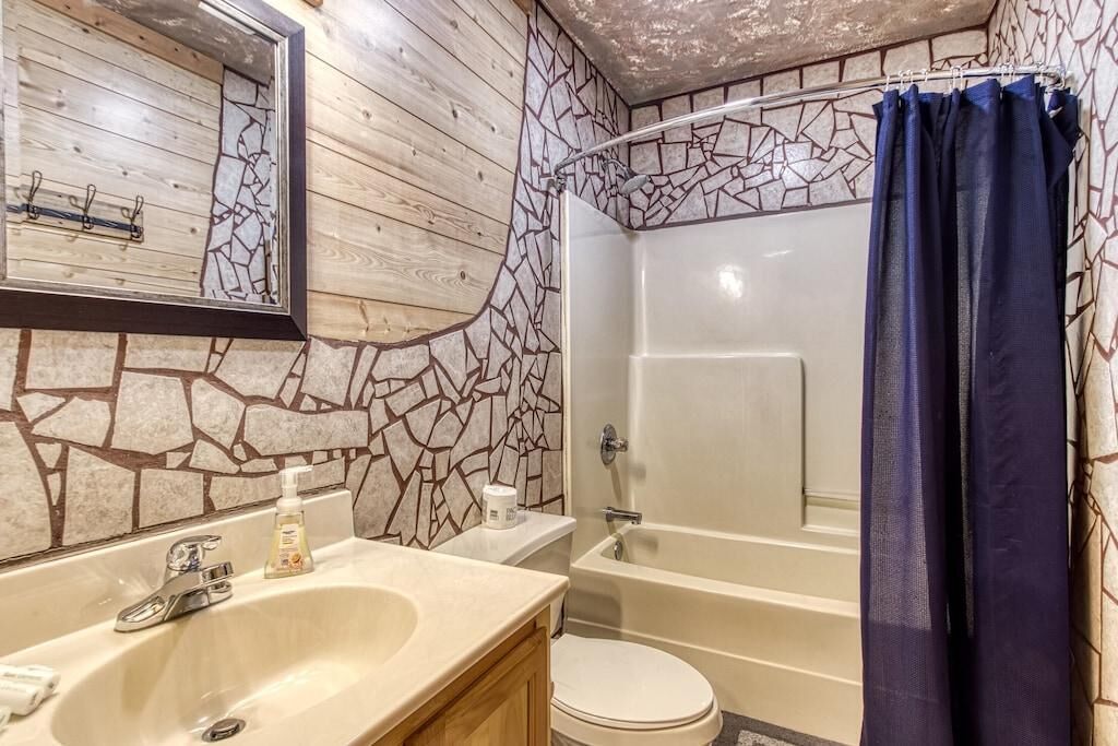 Un baño con pared de mosaico de piedra, tocador de madera, lavamanos, inodoro y bañera con cortina de ducha.