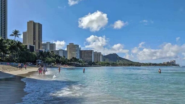 Une plage à l’eau cristalline, bordée de palmiers, avec des immeubles imposants en arrière-plan.