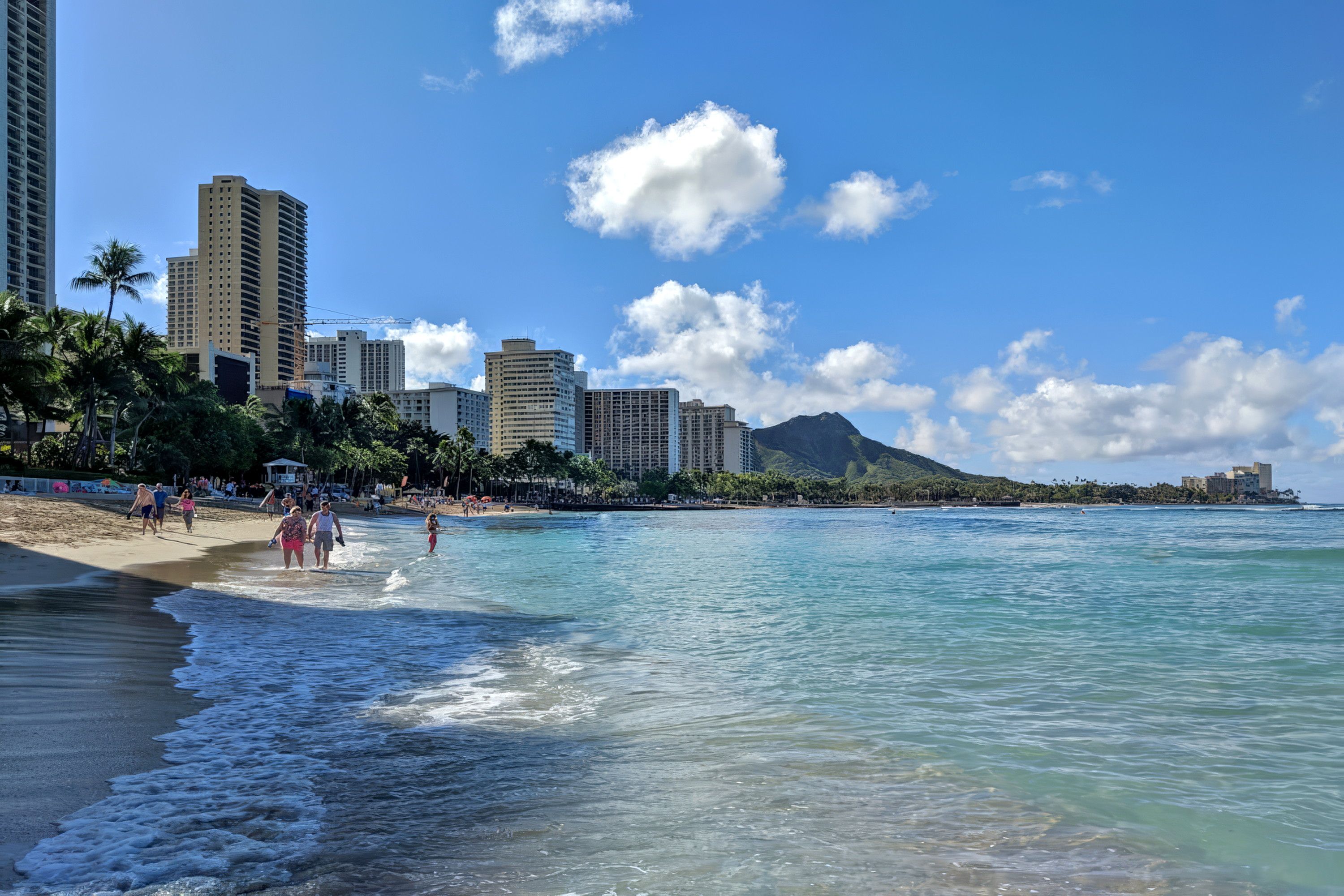 A beach with clear water, palm trees, and high-rise buildings in the background.